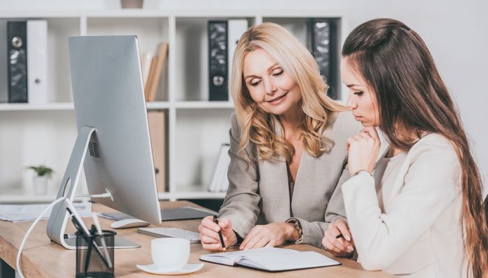 Smiling female business mentor pointing at notebook and working with young colleague in office